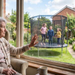 Elderly woman sitting in a chair and looking out the window at 3 children bouncing on a trampoline.